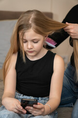 Young girl holding a smartphone, engrossed in the device while her mother is brushing her long blonde hair, depicting themes of family care and technology distraction. Vertical photo