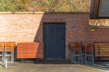 Stacked beer-garden tables and chairs rest by a brick wall. A dark wooden door sits in cool shadow.