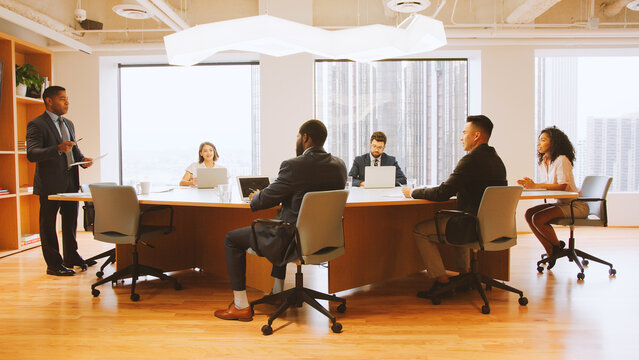 Mature Businessman Standing Giving Presentation To Colleagues Sitting Around Table In Modern Office