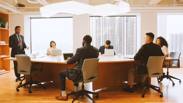 Mature Businessman Standing Giving Presentation To Colleagues Sitting Around Table In Modern Office - Powered by Adobe