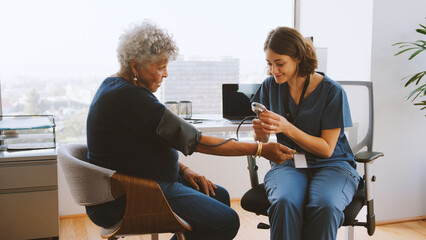 Nurse Wearing Scrubs In Hospital Office Checking Senior Female Patients Blood Pressure