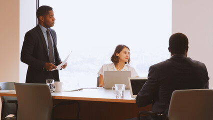Mature Businessman Standing Giving Presentation To Colleagues Sitting Around Table In Modern Office
