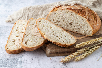 Sliced fresh baked spelt wheat loaf of bread on wooden cutting board with  rustic linen towel and spikelets on grunge stone table against white wall.