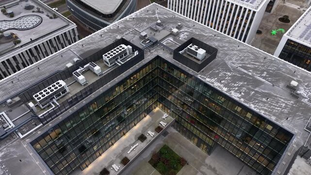 Aerial close-up of a glass-clad office building in Vilnius, Lithuania, showing illuminated workspaces and a rooftop with industrial HVAC systems and ventilation infrastructure.