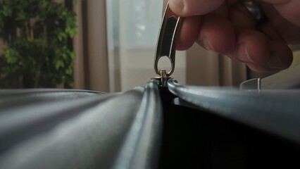 A Macro shot of a hand unzipping a suitcase with a plastic zipper and metal pull tab in a brightly lit room.