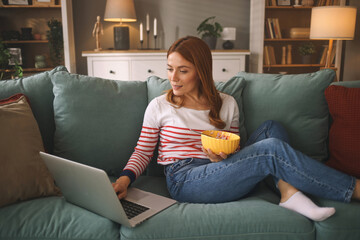 A woman sits comfortably on a couch with her legs crossed, holding a bowl of snacks. She is focused...