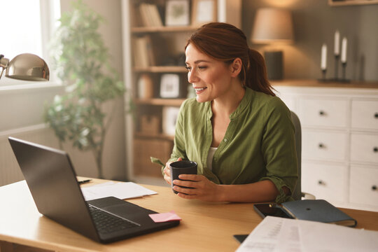 A woman sits at a wooden desk in a warm home office, engaging in a video chat with a laptop. She holds a cup of coffee and appears cheerful, surrounded by greenery and soft lighting. - Powered by Adobe