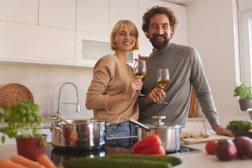 A couple stands in a modern kitchen, surrounded by fresh ingredients and cooking utensils. They are smiling and toasting with glasses of wine, showcasing a joyful cooking experience.