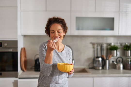 A woman with curly hair smiles while eating a bowl of cereal in a clean, modern kitchen. She is enjoying her breakfast with cheerful enthusiasm under natural light.