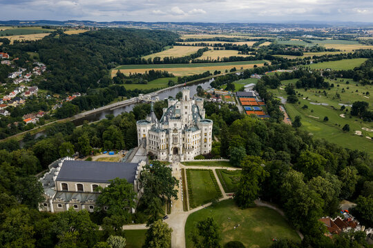 Aerial view of the majestic Zamek Hlubok&aacute; castle rises amidst lush greenery, with the Vltava River snaking through golden fields, Hluboka nad Vltavou, Jihocesky kraj, Czechia.
