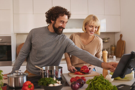 In a bright, modern kitchen, a man and woman share a joyful moment while cooking. They are chopping vegetables and following a recipe on a tablet, excitedly engaging with one another. - Powered by Adobe
