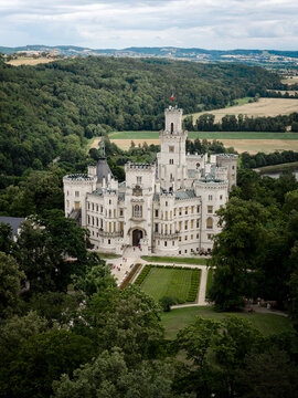 Aerial view of the majestic, ivory-white Hlubok&aacute; Castle, its spires piercing the sky amidst a sea of emerald forests and golden fields, Hluboka nad Vltavou, Jihocesky kraj, Czechia.