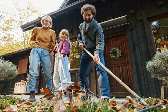 A family enjoys their weekend in autumn by raking leaves outside their cabin. They work together while laughing, surrounded by vibrant fall colors and a warm atmosphere. - Powered by Adobe
