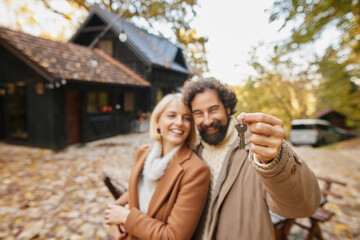 Fototapeta premium Happy couple showcases their new home keys while standing outside a charming cabin surrounded by colorful autumn leaves. Excitement fills the air as they start a new chapter.