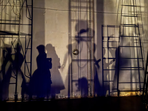 Shadows of ballet dancers and stage crew projected on a backstage wall under dramatic lighting, creating artistic silhouettes and geometric ladder patterns in a theater performance setting.