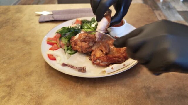 A man's hands in black latex gloves cut up baked chicken legs. The chicken is served on a plate with salad dressing and sauce. High quality 4k footage