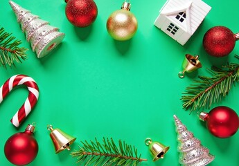 Flat lay of red and gold baubles, candy cane, glitter trees, white ceramic house, fir sprigs and tiny bells on green background