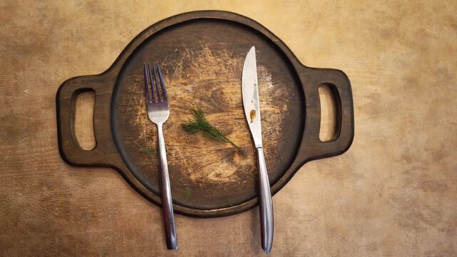 An empty vintage wooden plate with the remains of food and cutlery, a fork, and a knife. The food was eaten, leaving only a sprig of dill on the plate. High quality 4k footage