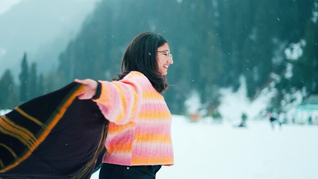 Indian woman smiling in the snowfall, happy twirling with wearing warm shawl. Tourist enjoying snowfall while standing in snow covered mountains at Gulmarg, Kashmir, India. Travel, holidays concept
