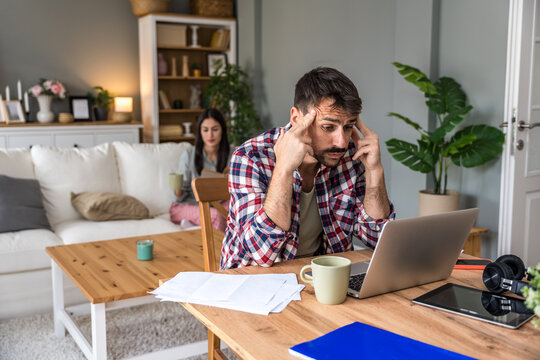 Man reviews bills at a laptop while his partner relaxes with a tablet on the sofa in a cozy living room. Papers, coffee and tech on the table. Concept: home budgeting, monthly expenses