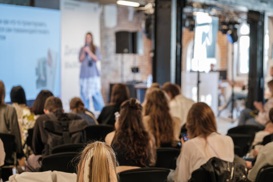 Conference hall with speaker on stage delivering presentation to attentive audience, attendees sit in rows under bright lights beside brick walls and screens.