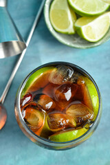 Glass of cold Cuba Libre cocktail and bowl with limes on blue background