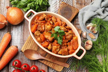 Delicious cooked stew in baking dish, fresh ingredients and spoon on wooden table, flat lay