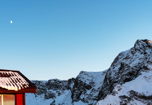 Snow-covered mountains under a clear blue sky with a small red house in the foreground. The moon is visible in the sky, adding to the serene winter landscape, copy space