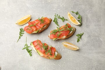 Tasty bruschettas with salmon, cream cheese, cucumber, arugula and lemon on grey table, flat lay