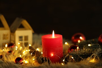 Burning candle, fir tree branches, cones, baubles and festive lights on faux fur against black background, closeup. Christmas greeting card