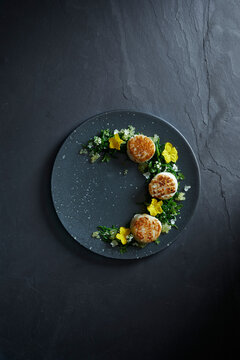 Gourmet food photography of seared scallops with herbs and edible flowers, plated on a stylish black stone plate for modern fine dining presentation