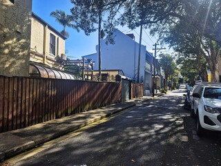Obraz premium Landscape view of an urban back street showing an arched tin awning behind a fence drenched in shadows from adjacent trees.