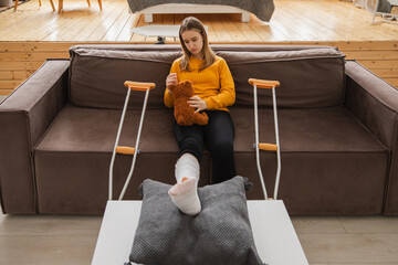 Young girl with broken leg cast sitting on sofa. Sad little kid hugging her teddy bear recovering from broken leg at home. Girl using cast and crutches. Accident injury fracture treatment foot bandage