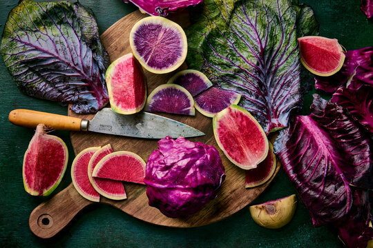 A vibrant still life featuring sliced watermelon radishes and red cabbage, arranged artfully on a wooden cutting board with a rustic knife, all set against a custom dark green background.