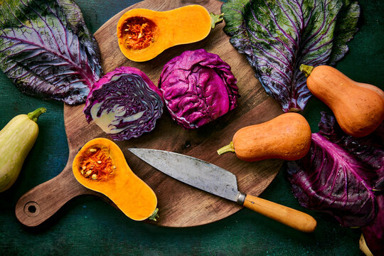 A vibrant still life featuring sliced butternut squash and red cabbage, arranged artfully on a wooden cutting board with a rustic knife, all set against a custom dark green background.