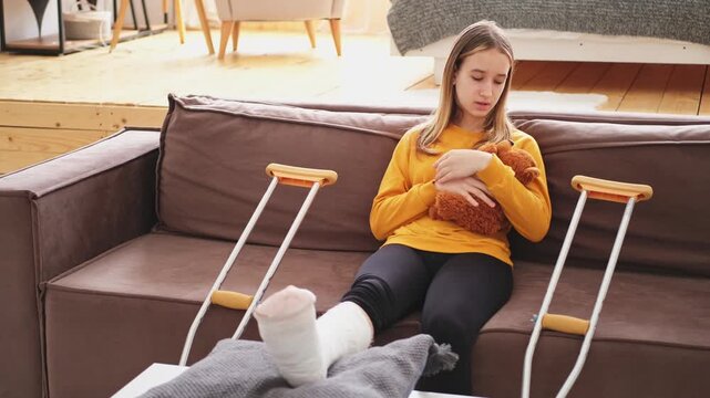 Young girl with broken leg cast sitting on sofa. Sad little kid hugging her teddy bear recovering from broken leg at home. Girl using cast and crutches. Accident injury fracture treatment foot bandage