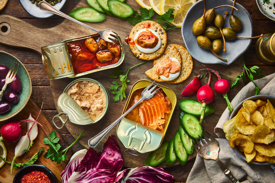 An overhead, flat lay of a luxurious tinned seafood appetizer or charcuterie board with tinned mussels, tuna and salmon,with sliced cucumbers, radishes, lemons, radicchio, olives,capers, potato chips