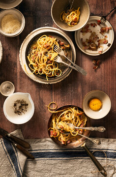 top view of wooden table set with bucatini carbonara in copper pan and plates and bowls accompanied with egg yolk, bacon and pepper in mortar, rustic messy scene