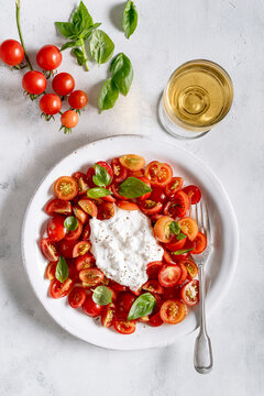 Plate with halved cherry tomatoes, topped with Italian stracciatella cheese and decorated with fresh bright green basil leaves, all on a light background..