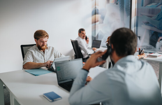 A bustling office scene with colleagues using laptops, tablets, and phones.