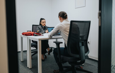 Two colleagues sit across from each other at a bright, white desk in a modern office, collaborating over a laptop and papers, conveying focus, communication, and teamwork in a professional setting.