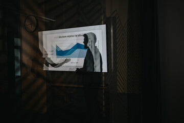 Two professionals exchange a handshake as a chart is shown on a large screen. The scenes reflections and shadows create a moody, collaborative atmosphere focused on strategy and teamwork.