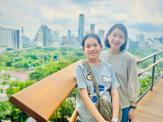 Asian preteen girl and middle-aged mother stand on a rooftop garden in Bangkok, smiling. The city skyline with tall buildings and lush greenery is visible in the background, under a partly cloudy sky
