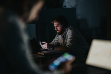A bearded man wearing headphones works intently at a desk with a laptop and phone in low light. A blurred colleague in the foreground suggests remote collaboration during late hours.