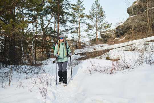 Caucasian young adult woman hiking through snowy forest trail carrying backpack and trekking poles, wearing sunglasses and winter gear, surrounded by pine trees and rocky landscape - Powered by Adobe