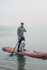 young man with tattooed arms on a sup board with his samoyed dog at the lake during sunset