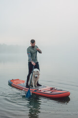 young man with tattooed arms on a sup board with his samoyed dog at the lake during sunset