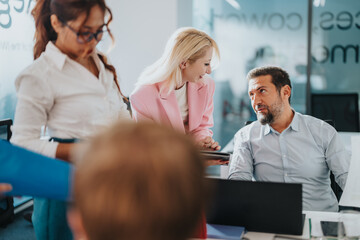 A diverse group of professionals collaborates around a desk as a woman in a pink blazer shares...