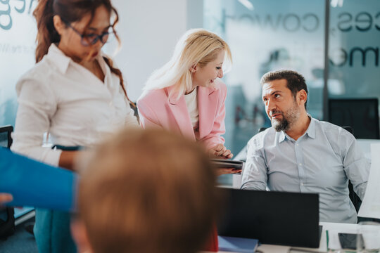 A diverse group of professionals collaborates around a desk as a woman in a pink blazer shares ideas with a man in a light shirt. They review documents and devices in a bright office.