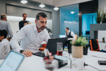 A focused professional sits at a bright office desk, typing on a laptop as teammates work nearby....